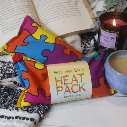 Two Mini Heat packs are stacked upon each other, one with a belly band label reading HEAT PACK LUPIN FIlled.   These are resting on a black and white blanket with open book, candle and coffee cup evoking a cosy feel.    The Heat packs feature brightly coloured jigsaw puzzle pieces connected together