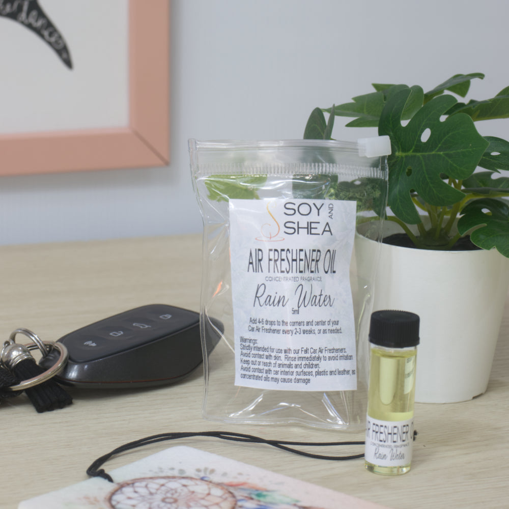 A small clear bottle with black cap of Soy's Shea Air Freshener Oil next to a clear resealable pouch, displayed on a light wood tabletop with a car key, a felt air freshener, and a potted plant in the background.  