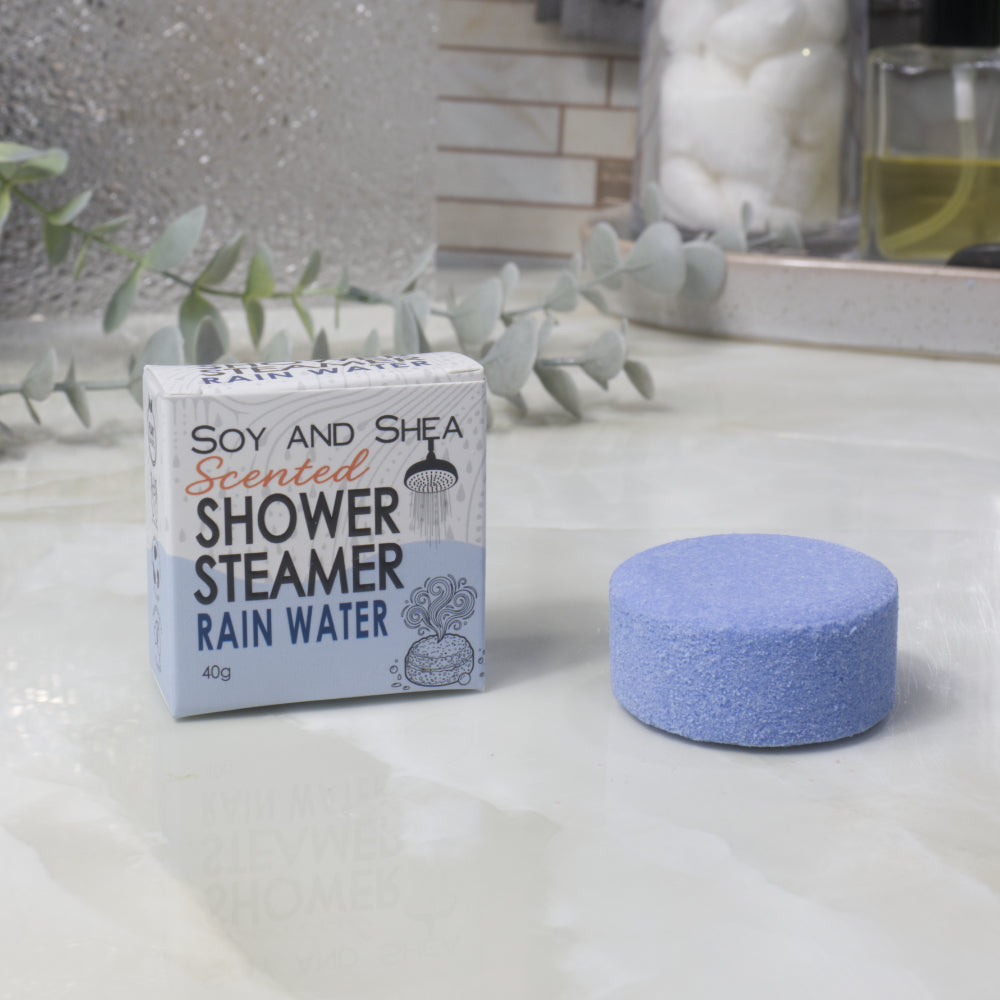 A boxed scented shower steamer and the steamer itself on a bathroom counter. The box reads 'Soy and Shea Scented Shower Steamer Rain Water', and the round blue-purple steamer sits beside it.