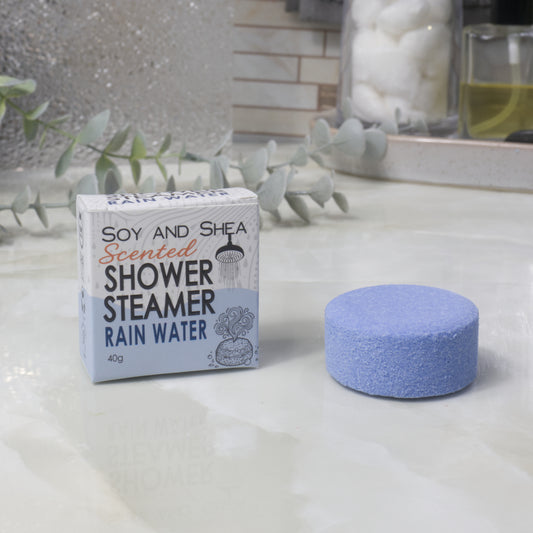 A boxed scented shower steamer and the steamer itself on a bathroom counter. The box reads 'Soy and Shea Scented Shower Steamer Rain Water', and the round blue-purple steamer sits beside it.