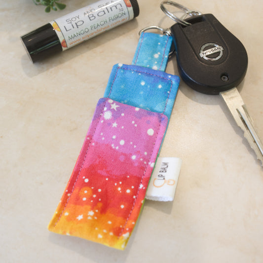 A clip balm holder lies on a wooden surface attached to a car key with a "lobster claw" clip.  To the side is a tube of lip balm. The Clip Balm has layers of bright watercolour in a rainbow look with white dots and stars.