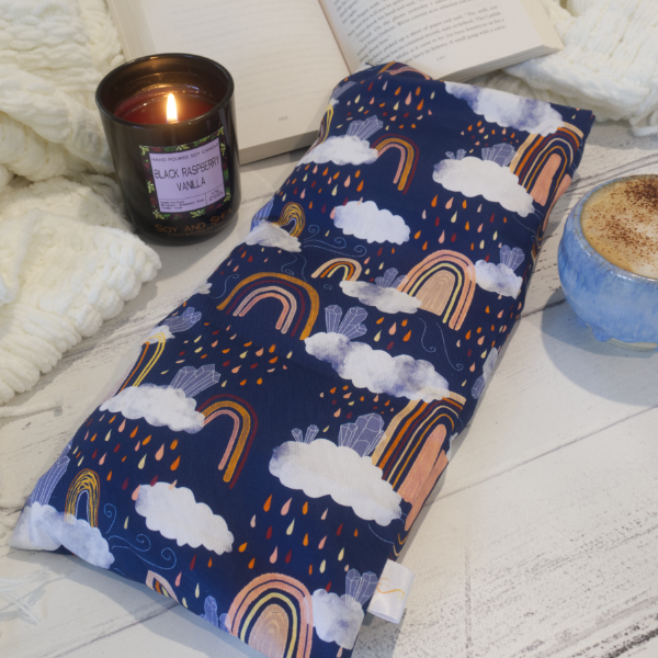 A long rectangular heat pack lays on a wooden table resting against an open book upon a white blanket. To the side is a burning candle and a cup of coffee.  The Heat Pack is dark blue with drawings of Boho style rainbows in shades of orange and yellow surrounded by white clouds with coloured raindrops.  Upon the clouds sits clusters of blue crystals