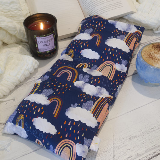A long rectangular heat pack lays on a wooden table resting against an open book upon a white blanket. To the side is a burning candle and a cup of coffee.  The Heat Pack is dark blue with drawings of Boho style rainbows in shades of orange and yellow surrounded by white clouds with coloured raindrops.  Upon the clouds sits clusters of blue crystals