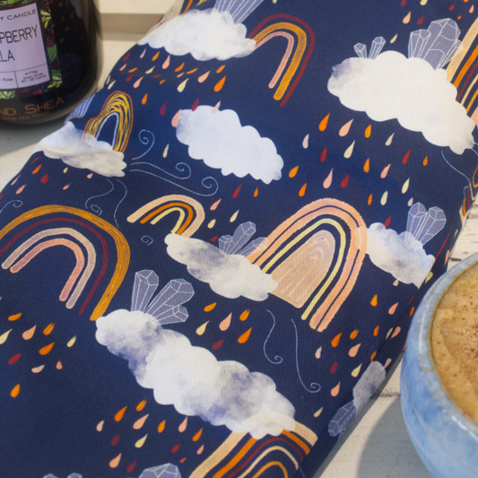 A close up image of the Rainbows Crystals Heat Pack focusing on the fabric design.  The heat pack is a dark blue fabric featuring boho style rainbows in shades of orange.  These are surrounded by rain clouds with water drops falling in shades of orange, red and yellow and a cluster of crystals sit on each cloud.