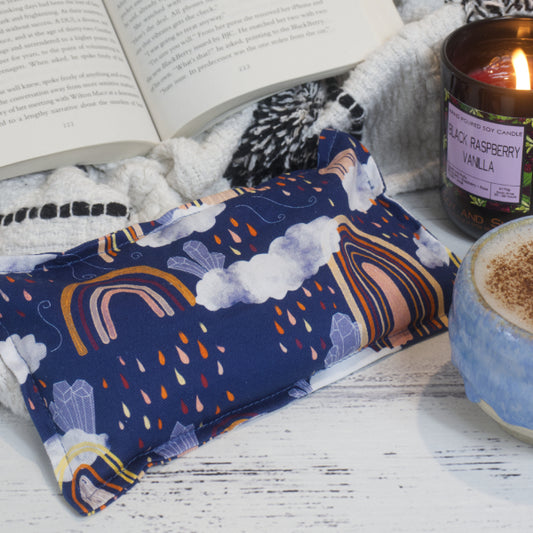 A mini heat pack with a multicolored pattern, including rainbows, rain clouds and crystals displayed on a wooden surface next to an open book, a plush blanket, lit candle, and a cup with a beverage.