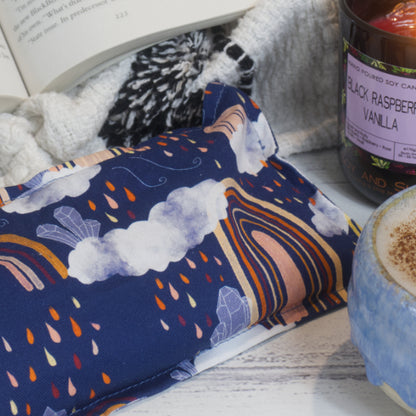 A close up of the Rainbow Crystals Mini Heat pack showing the fabric pattern. It has a dark blue backgroudn with orange boho rainbows, rain clouds with rain and crystals,