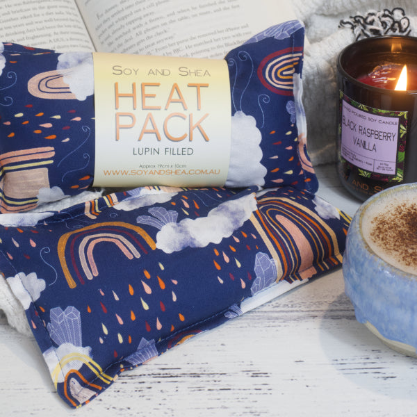 Two Mini rectangular heat packs sit on a bench featuring a multicolored pattern, including rainbows, rain clouds and crystals.  These are next to an open book, a plush blanket, lit candle, and a cup with a beverage.