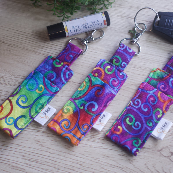 A selection of three clip balms showing variations in fabric design, lie on a wooden side with the middle Clip Balm attached to a car key.  A lip balm lays near-by along with some greenery.  The Clip balms have a deep rich rainbow tie dye background with bright rainbow filigree like swirls over it.  Each Lip Balm Holder shows a slight variation in fabric design.