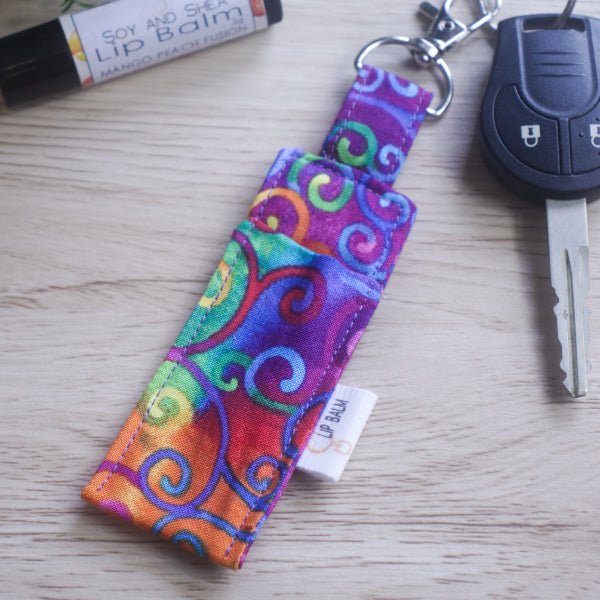 A lip balm holder lies on a wooden surface attached to a car key with a "lobster claw" clip.  To the side is a tube of lip balm. The Clip Balm has a dark rainbow tie dyed backgound with filigree rainbow swirls.