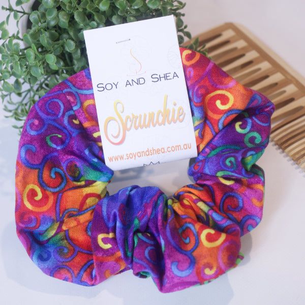 A colorful scrunchie sits on wooden surface resting against a wooden comb with greenery in the background.  A cardboard tag reads SOY AND SHEA SCRUNCHIE.    The scrunchie has a rich tie dyed background covered in rainbow filigreen like swirl swirls.