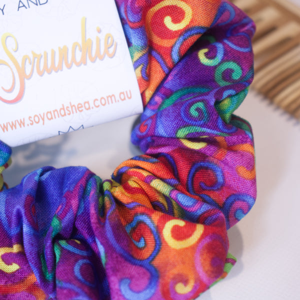 A close up of the fabric design on the Rainbow Curls scrunchie showing the rich tie dye rainbow background with colourful filigree swirls.