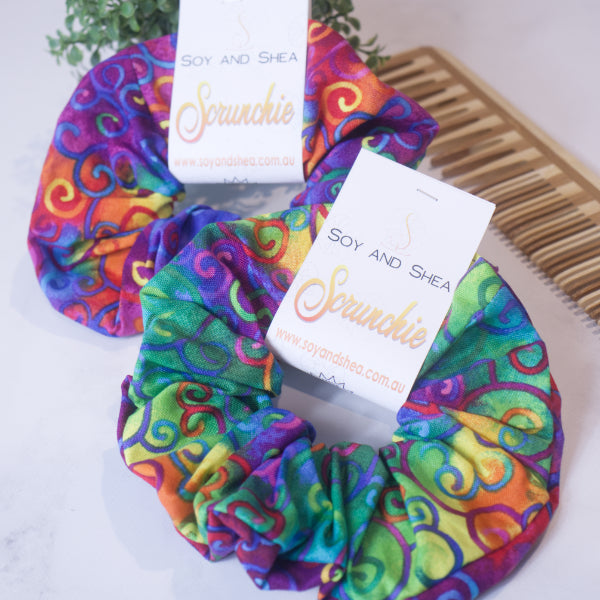 Two colourful scrunchies displayed on a white surface next to a wood comb and greenery.  The scrunchies feature a rich rainbow tie dye fabric witlh colourful filigree swirls.  Each Scrunchie shows a slight variation in the fabric design.