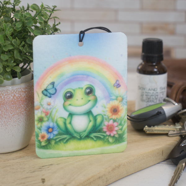 A car air freshener featuring an illustration of a frog with a rainbow and flowers in the background, rests against a pot plant next to a set of car keys