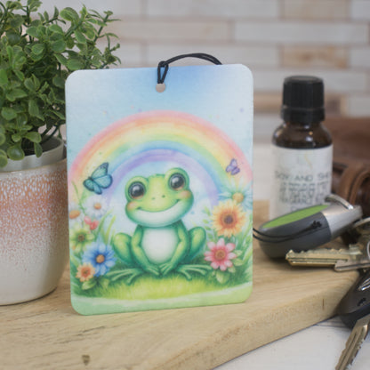A car air freshener featuring an illustration of a frog with a rainbow and flowers in the background, rests against a pot plant next to a set of car keys