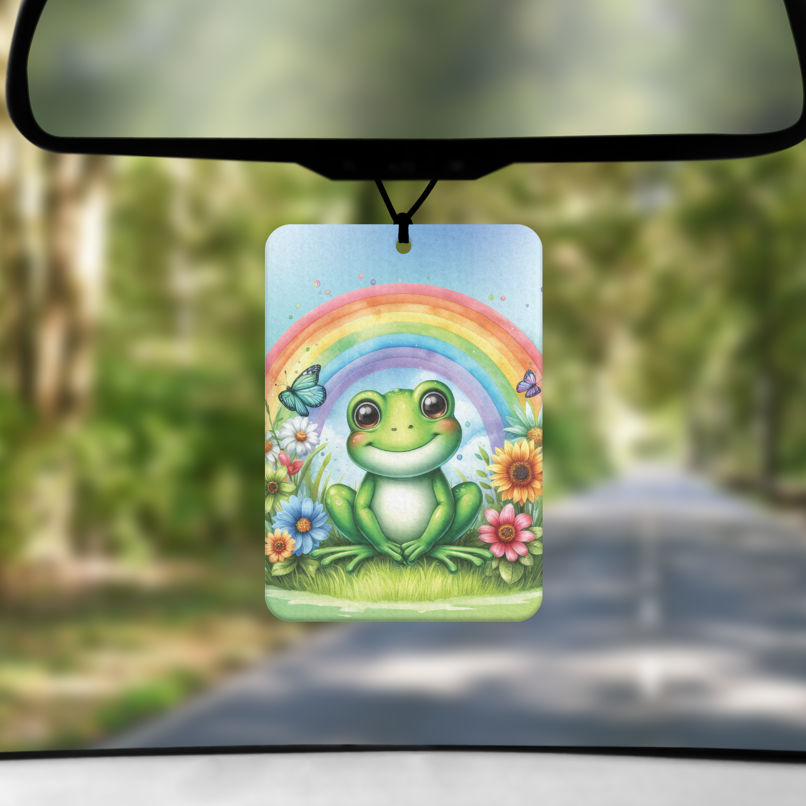 A mock up image showing a view through a car windscreen with a long tree lined road.  From the rearview mirror hands an air freshener with an image of green frog sitting under a rainbow and surrounded by flowers