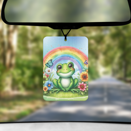 A mock up image showing a view through a car windscreen with a long tree lined road.  From the rearview mirror hands an air freshener with an image of green frog sitting under a rainbow and surrounded by flowers