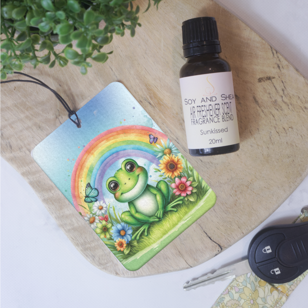 A car air freshener featuring an illustration of a frog with a rainbow and flowers in the background, lies on a wooden block next to a bottle of Air Freshener scent