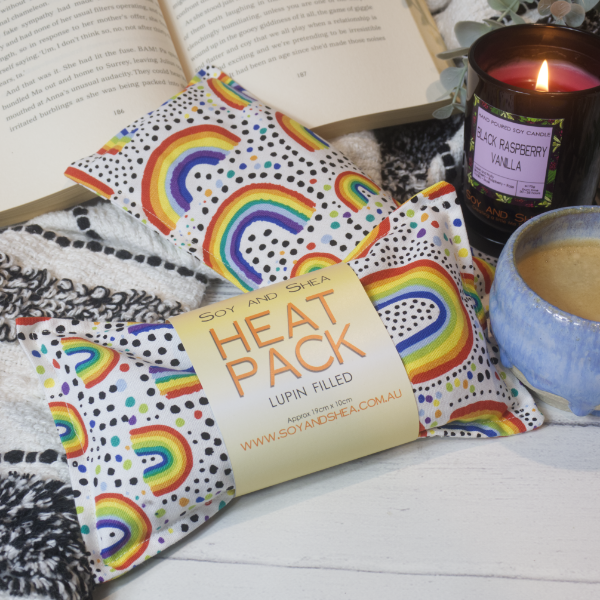Two Mini Heat packs are stacked upon each other, one with a belly band label reading HEAT PACK LUPIN FIlled.   These are resting on a black and white blanket with open book, candle and coffee cup evoking a cosy feel.    The Heat packs are white with drawings of various sized rainbows surrounded by dots in rainbow and black colours