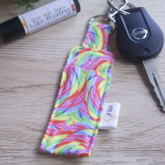 A lip balm holder lies on a wooden surface attached to a car key with a "lobster claw" clip.  To the side is a tube of lip balm. The Clip Balm has brignt rainbow coloured round swirls in the style of rainbow lollipops