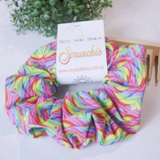 A colorful scrunchie sits on wooden surface resting against a wooden comb with greenery in the background.  A cardboard tag reads SOY AND SHEA SCRUNCHIE.    The scrunchie has a colourful rainbow striped pattern with white stripes separating the colours