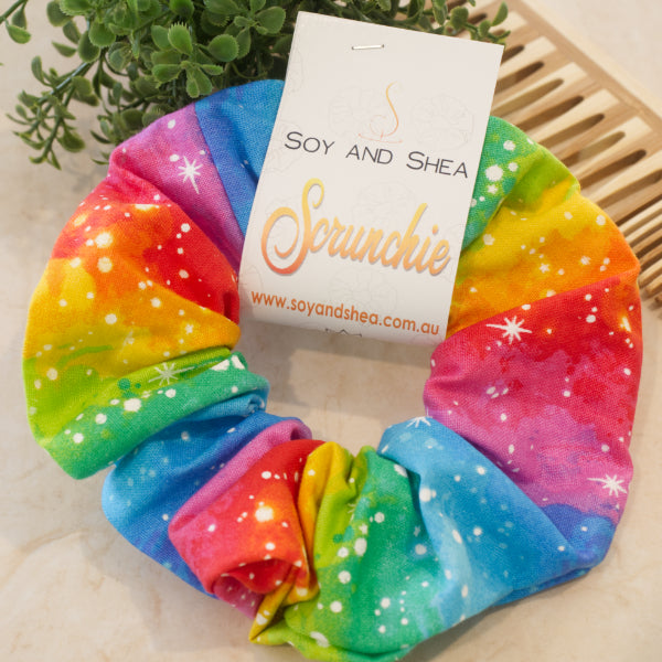 A colourful scrunchie sits on a white surface next to a wooden comb and green plant. The scrunchie features rows of bright rainbow colours in a watercolour effect with white dots and stars.
