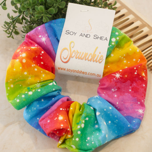 A colourful scrunchie sits on a white surface next to a wooden comb and green plant. The scrunchie features rows of bright rainbow colours in a watercolour effect with white dots and stars.