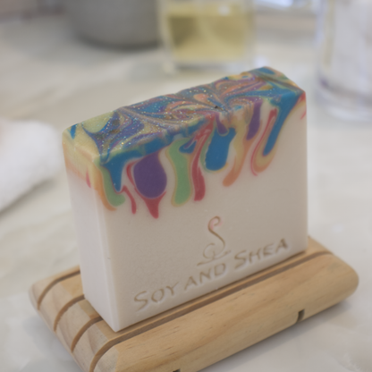 A side shot of a Rainbow Sparkle Soap bar on a wooden soap dish with a blurred background of a bathroom counter-top.  The soap has a cream base layer with drips of rainbow colours coming down from the top.
