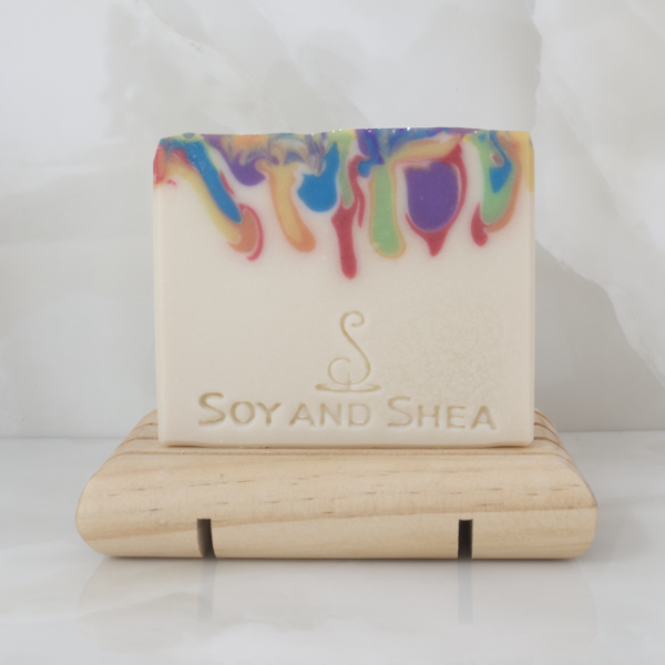 A soap with a cream base layer and drips of rainbow colours coming down from the top sits on a wooden soap dish on a tiled background