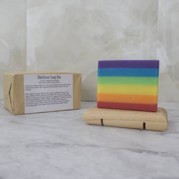 A rainbow layered soap bar sits on a wooden stand with a brown paper package behind labeled 'Rainbow Soap Bar' on a marble background
