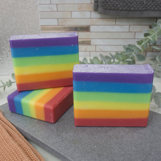 Three rainbow-colored soap bars on a stone surface with a tiled wall and plant in the background.  The soaps feature six even layers of rainbow colour starting with red at the bottom and finishing with purple