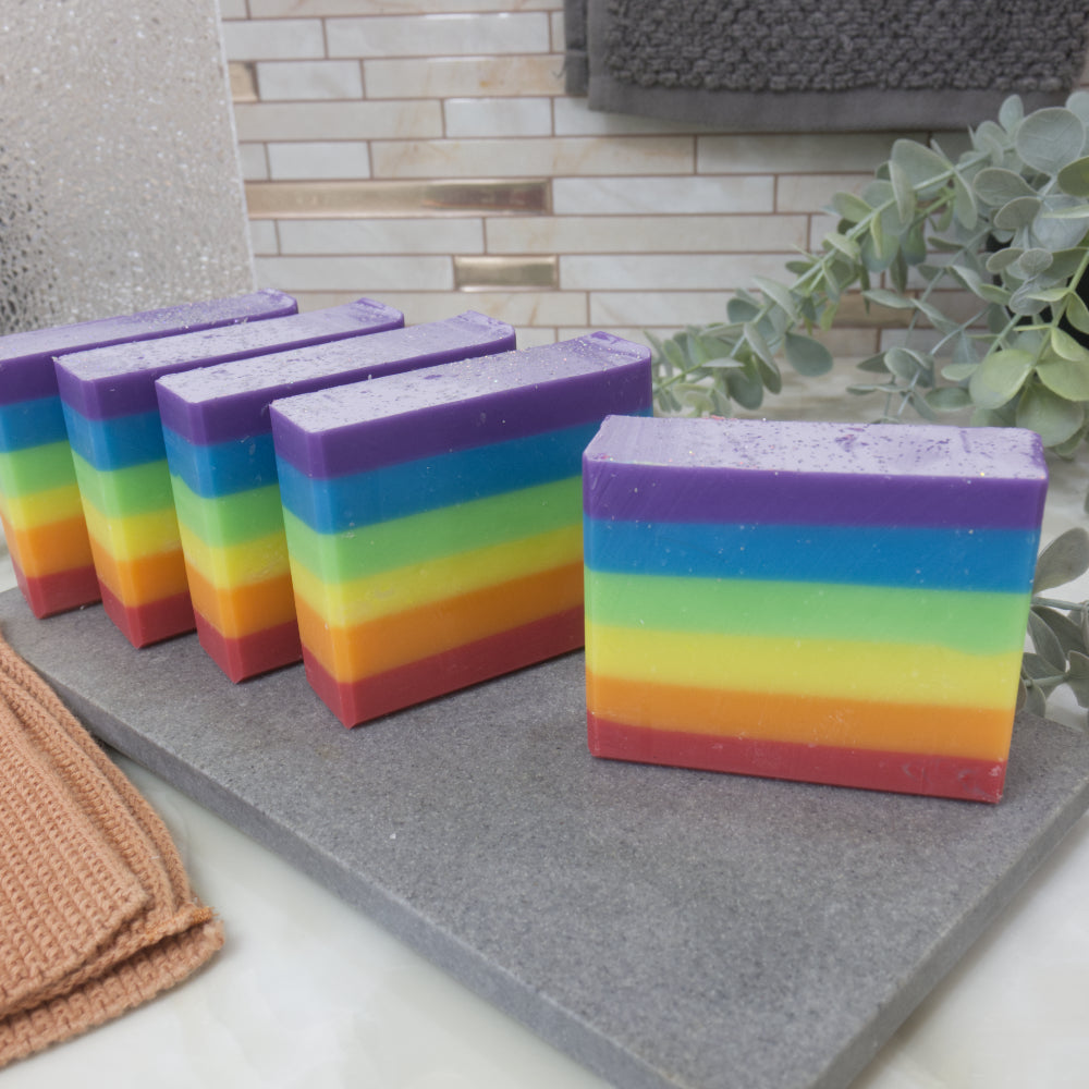 Rainbow striped soap bars on a gray surface with a tiled wall and plant in the background.  The soaps feature six even layers of rainbow colour starting with red at the bottom and finishing with purple