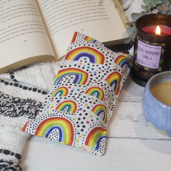 A mini rectangular heat pack rests against a black and white blanket and open book.  Next to it is a burning candle and filled coffee cup evoking a comfy scene.   The Heat Pack is a white fabric with various sized stylised rainbows scattered over it surrounds by dots in rainbow and black colours