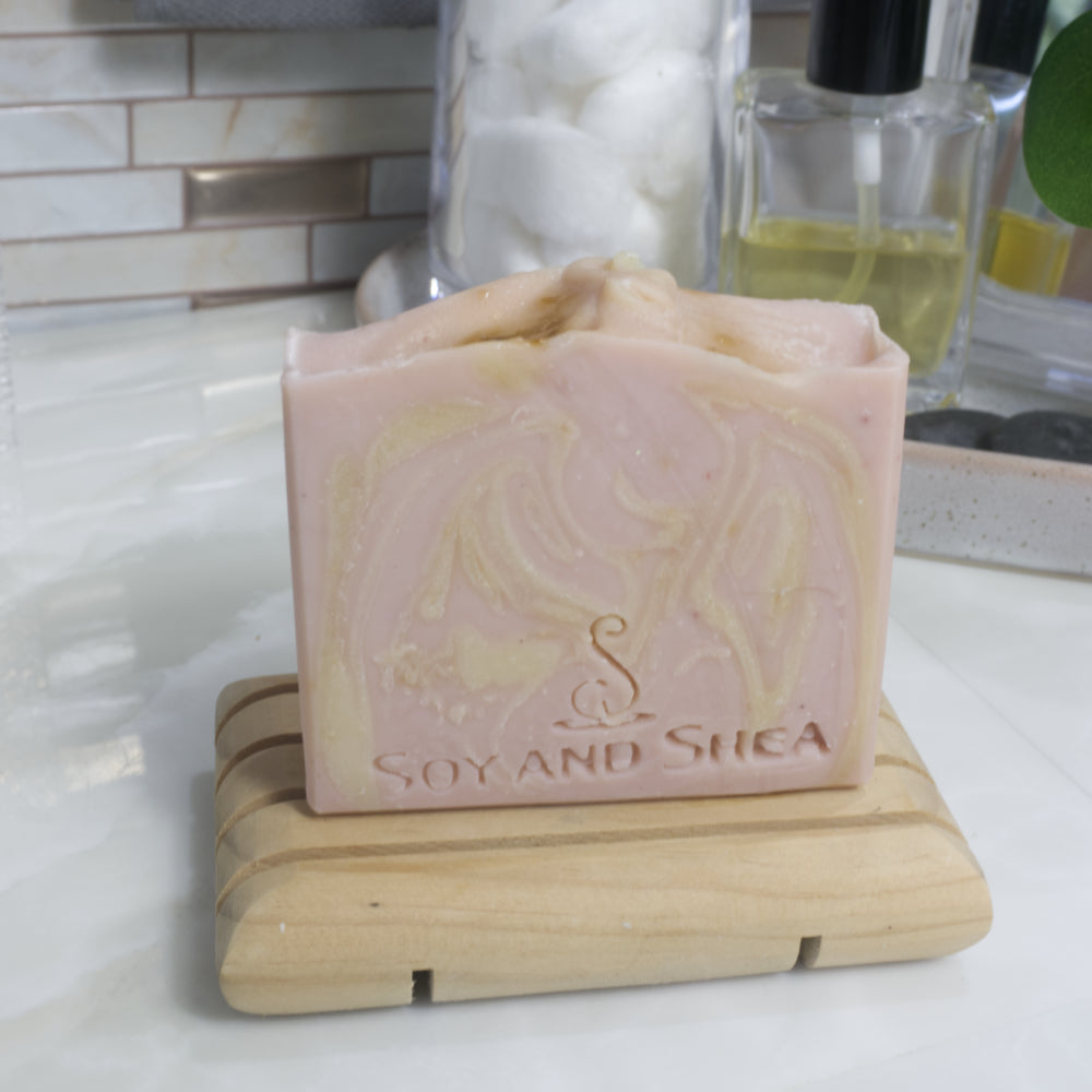 A pale pink soap bar with cream swirls stands on a wooden soap rack on a bathroom countertop with a blurred background