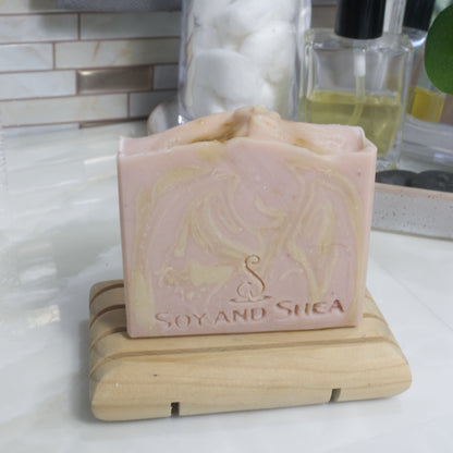 A pale pink soap bar with cream swirls stands on a wooden soap rack on a bathroom countertop with a blurred background