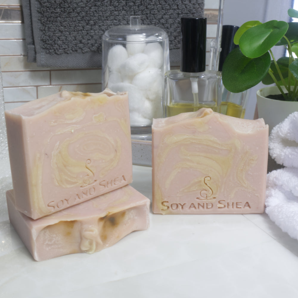 A select of three soap are displayed on a bathroom countertop with a white towel, perfume and jar of cotton balls. To the front left a soap bar lays flat revealing the wavy top, while another stands upright on it.  To the right, a third soap stands upright.  All soaps have a pale pink base colour, with creamy-yellow swirls throughout