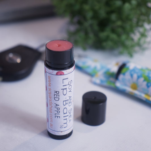 A black tube with colourful label reading LIP BALM RED APPLE stands upright on a white counter with a blurred background.  The black cap has been removed and sits to the side revealing a red balm