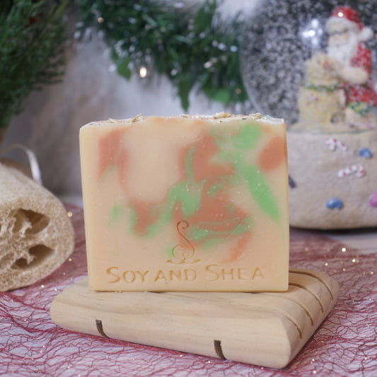 A vibrant Reindeer Food Soap Bar from Soy and Shea, infused with peppermint and rosemary essential oils, rests on a wooden soap dish. The scene is set with festive holiday decorations featuring a tiny Santa figurine and lush green foliage, offering a warm, handmade soap experience.