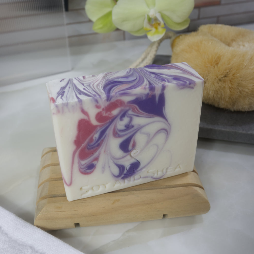 White soap bar with pink and purple swirls on a wooden soap dish, with a blurred background.