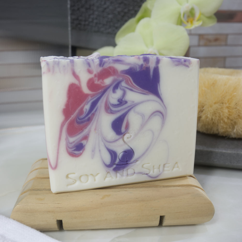 White soap bar with pink and purple swirls on a wooden soap dish, with a blurred background.
