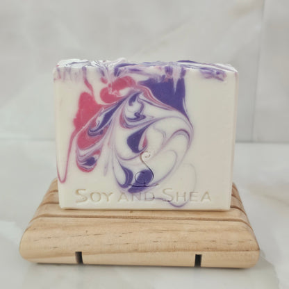 White Soy and Shea soap bar with pink and purple swirls sits on a wooden soap dish against a white background