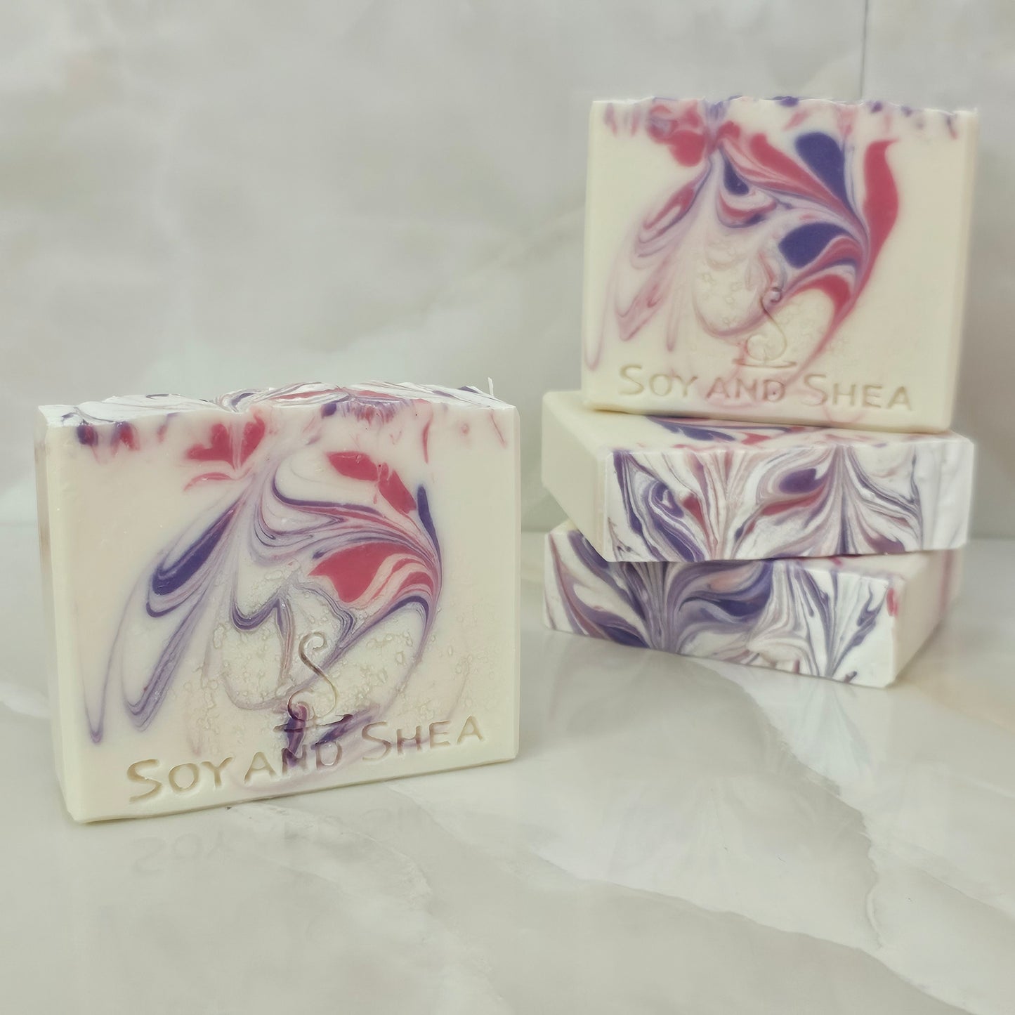 A selection of Soy and Shea soap bars that are white with pink and purple swirls sit on marble tiles