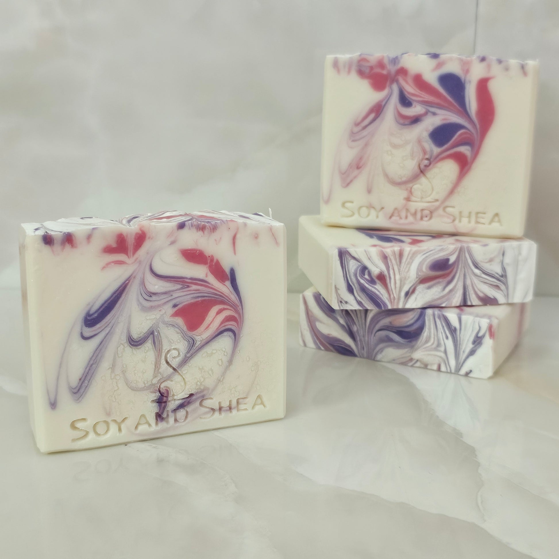 A selection of Soy and Shea soap bars that are white with pink and purple swirls sit on marble tiles