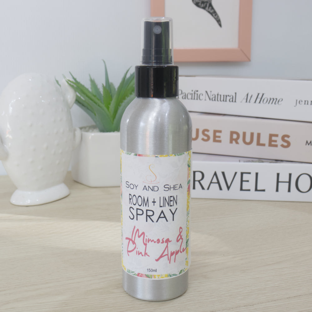 A silver aluminum room and linen spray bottle with a black spray top, standing on a light wood surface. The bottle has a white label with colorful floral accents and black text reading 'SOY AND SHEA ROOM + LINEN SPRAY' with 'Mimosa & Pink Apple' in pink script.