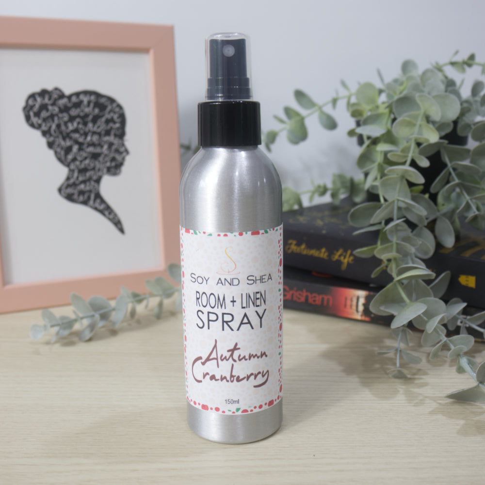 An aluminium bottle with black spray cap stands on a wooden surface surround by a pile of books with a plant sitting on top of them and a picture frame to the left.   The bottle features a decorative product label identifying it as Room + Linen Spray, scented in Autumn Cranberry