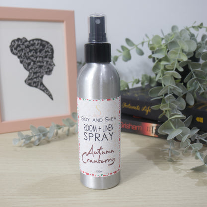 An aluminium bottle with black spray cap stands on a wooden surface surround by a pile of books with a plant sitting on top of them and a picture frame to the left.   The bottle features a decorative product label identifying it as Room + Linen Spray, scented in Autumn Cranberry