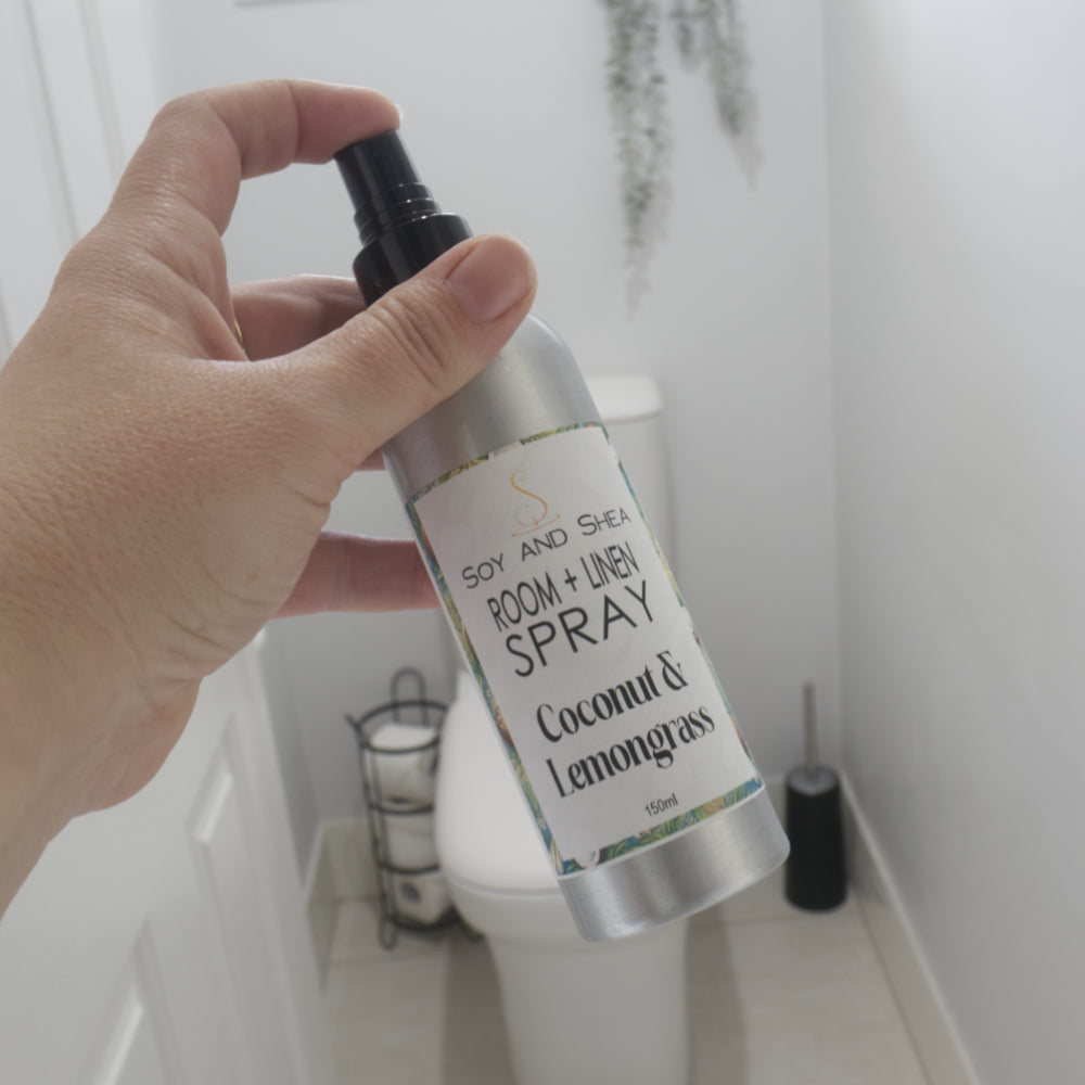 A hand holds a silver room spray bottle labeled 'Soy and Shea Room + Linen Spray' in a bathroom setting with a toilet visible in the background.  It is labelled as Coconut & Lemongrass