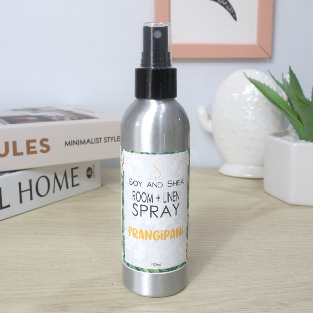 A silver metal spray bottle of Soy and Shea Room + Linen Spray with a black spray cap, standing on a light wood desk with a stack of books and a small potted plant in the background.  The label shows this has scented in Frangipani