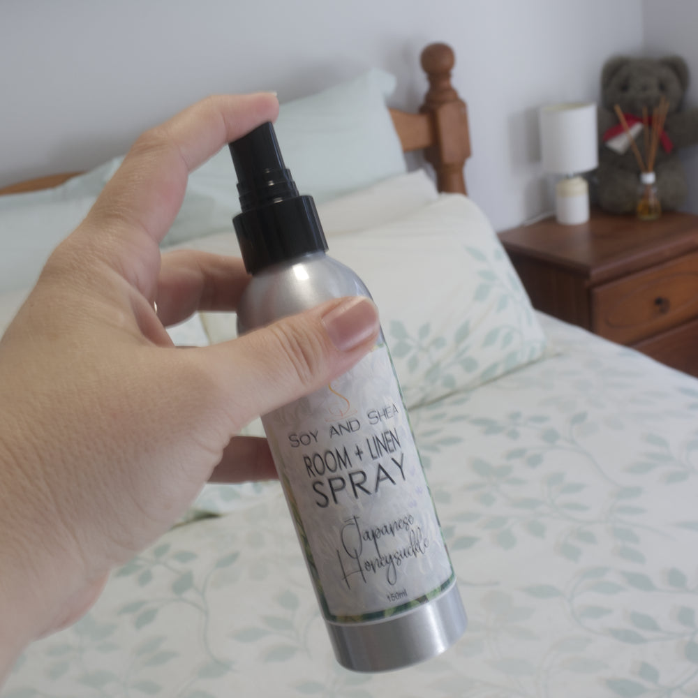 A hand holding a silver room and linen spray bottle with a black spray top. The label reads 'Soy and Shea ROOM + LINEN SPRAY Japanese Honeysuckle'.  The background is a blurred setting of a bed with side table with lamp