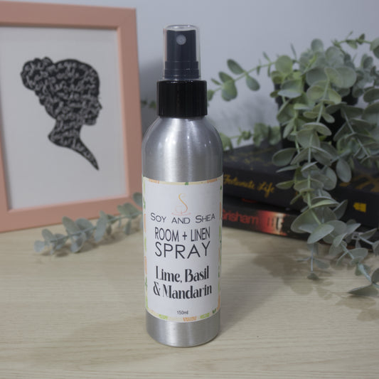 An aluminium bottle with black spray cap stands on a wooden surface surround by a pile of books with a plant sitting on top of them and a picture frame to the left.   The bottle features a decorative product label identifying it as Room + Linen Spray, scented in Lime, Basil and Mandarin