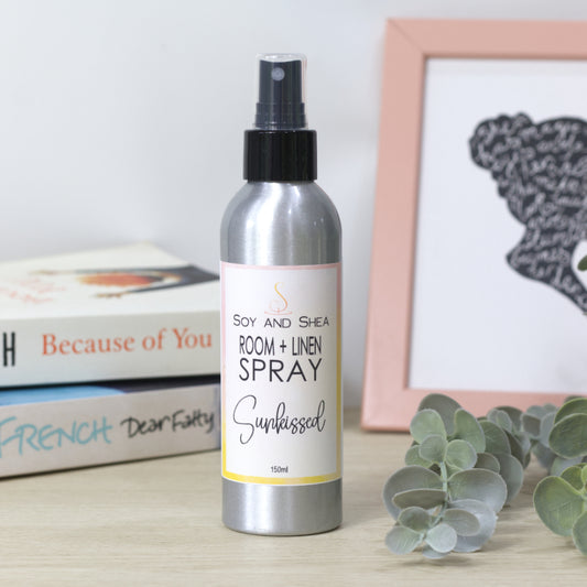 A 150ml silver metal spray bottle of Soy and Shea Room + Linen Spray, labeled 'Sunkissed', standing on a light wooden surface next to books, a framed picture, and eucalyptus leaves.